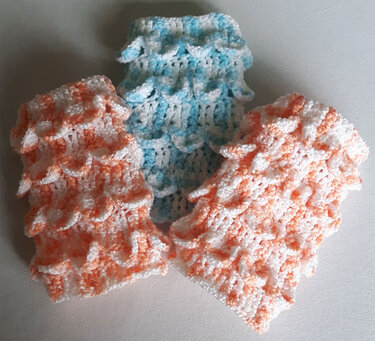 Drei gehäkelte Stulpen in Pastellfarben (Orange/Weiß, Hellblau/Weiß) mit schuppenartigem Muster auf hellem Untergrund