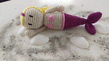 Gehäkelte Amigurumi-Meerjungfrau mit gelben Haaren, rosa Bikinioberteil und lila Schwanz auf Sand neben Muscheln