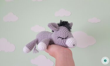 hellgrauer gehäkelter Amigurumi-Esel mit dunkler Mähne und weißen Akzenten, schlafend in einer Hand vor hellgrünem Hintergrund mit Wolken