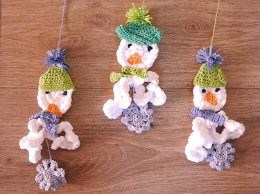 Drei gehäkelte Schneemann-Anhänger mit grünen Mützen und grauen Häkelblüten auf Holz