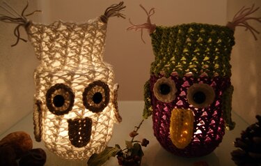 Zwei gehäkelte Eulen-Windlichter, ein weißes leuchtendes und ein violett-grünes Modell mit großen Augen