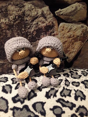 Zwei gehäkelte Amigurumi-Figuren mit grauen Kapuzen und kleinen schwarzen Gitarren auf einer Decke mit Leopardenmuster vor einer Steinwand.