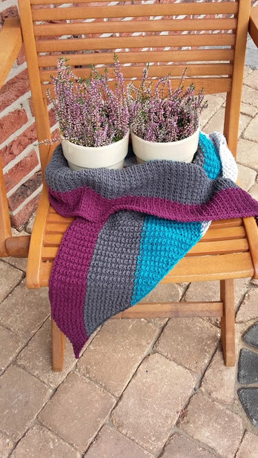 Gestricktes Tuch in Grau, Petrol und Violett auf Holzstuhl mit blühendem Heidekraut