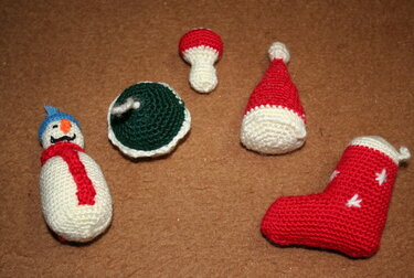 Fünf gehäkelte Weihnachtsfiguren auf braunem Untergrund: Schneemann, grüne Kugel, Fliegenpilz, rote Mütze und rote Socke.