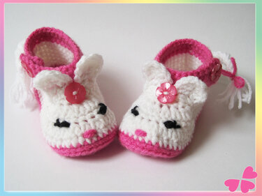 Zwei gehäkelte Babyschuhe in Weiß und Rosa mit Tiergesicht, Ohren, Blütenknopf und Riemen, von oben aufgenommen.