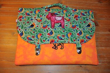 Orange Stofftasche mit grünem Überschlag mit Vogel- und Blattmuster und rosa Pferdeapplikation auf Holzoberfläche.