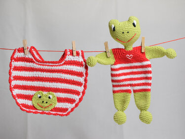 Gehäkeltes Lätzchen in Rot-Weiß mit grünem Froschgesicht und passende gehäkelte Frosch-Puppe, an einer roten Leine mit Wäscheklammern vor hellem Hintergrund.