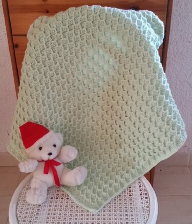 Pastellgrüne gehäkelte Babydecke mit Wabenstruktur auf Stuhl, weißer Teddybär mit roter Mütze davor