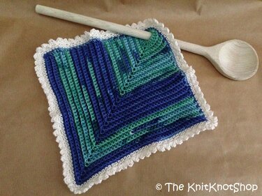 Quadratischer gehäkelter Topflappen in Blau und Grün mit weißem Rüschenrand neben einem Holzkochlöffel