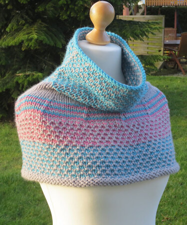 Gestrickter Damenponcho mit Rollkragen und Streifen in Grau, Rosa und Türkis auf einer Schneiderpuppe im Garten.