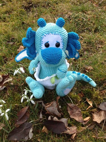 Gehäkelter türkis-blauer Amigurumi-Drache mit weißem Bauch sitzt im Gras neben Schneeglöckchen