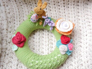 Grüner gehäkelter Kranz auf weißer, grob gestrickter Decke, dekoriert mit braunem Häkelhasen, gestreiftem Osterei und bunten Häkelblüten.