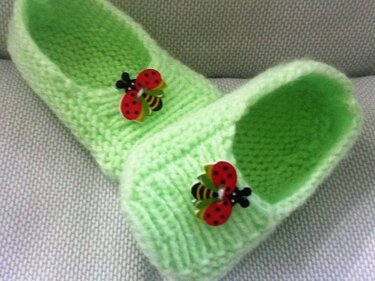 Paar hellgrüner gestrickter Babyschuhe mit roten Marienkäferknöpfen auf hellgrauem Untergrund