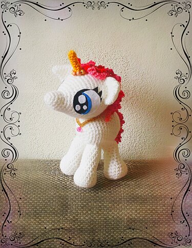 Weißes gehäkeltes Einhorn-Amigurumi mit pinker Mähne, gelbem Horn und goldener Halskette vor neutralem Hintergrund.