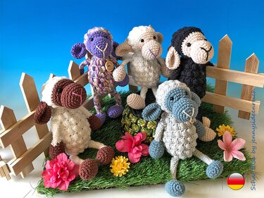 Fünf gehäkelte Amigurumi-Schafe in verschiedenen Farben auf Kunstgras mit Holzzaun und Blumen