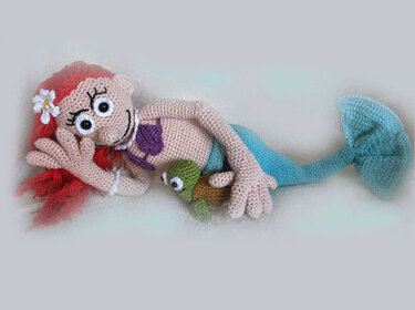 Hand-crocheted mermaid doll with red yarn hair, white hair flower, purple top and blue tail, posed beside a small green crocheted turtle on a neutral background.