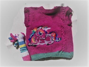 Magentafarbener gestrickter Pullunder mit buntem Einhorn- und Regenbogenmotiv, daneben ein gehäkeltes Einhornspielzeug.