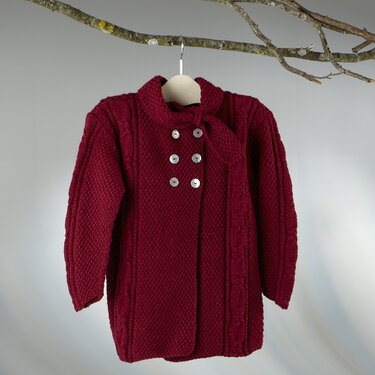 Dunkelrote gestrickte Kinderjacke mit sechs hellen Knöpfen und Schleife am Kragen, aufgehängt an einem Ast.