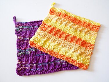 Zwei handgestrickte Topflappen, einer gelb-orange gestreift, der andere lila mit bunten Akzenten.