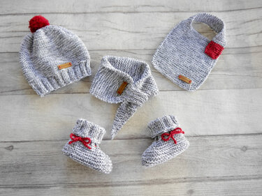 Flat-lay of a grey marled knitted baby hat with red pom-pom, triangular bandana scarf, square bib with red patch and pair of booties with red ties on a wooden surface