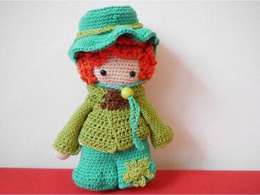 Gehäkelte Amigurumi-Puppe mit rostroten Locken, grünem Hut, grüner Jacke und türkisfarbener Hose mit Kleeblatt-Applikation