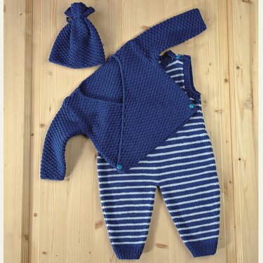 Marineblaues gestricktes Baby-Set mit Jacke, gestreiftem Overall und Mütze auf Holzuntergrund.