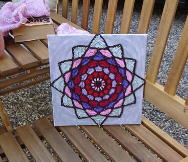 Gehäkeltes Mandala in Rot, Rosa, Violett und Hellblau auf quadratischer Leinwand auf einer Holzbank mit Wollknäuel