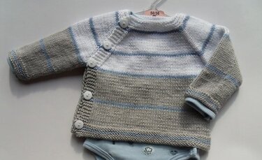 Gestrickte Babyjacke in Grau, Weiß und Hellblau mit seitlicher Knopfleiste auf einem Bügel