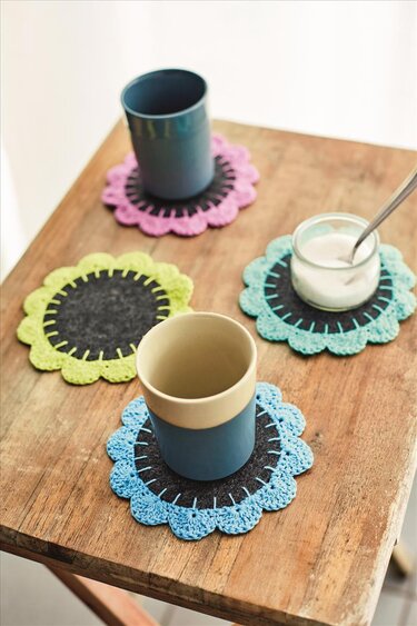 Vier gehäkelte, blumenförmige Untersetzer in Rosa, Grün und Blau auf einem Holztisch, drei davon mit Tassen und einem Joghurtglas.