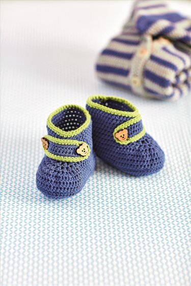 Gehäkelte dunkelblaue Babyschuhe mit hellgrünem Rand und kleinen Bärenknöpfen, auf gemustertem Untergrund mit gestreifter Decke im Hintergrund