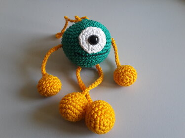 Gehäkelter einäugiger Amigurumi-Anhänger, grüner Körper, weißes Auge mit schwarzem Knopf und gelbe Kugelanhänger