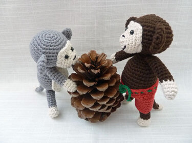 Zwei stehende gehäkelte Amigurumi-Affen (grau und braun) mit Kiefernzapfen auf hellem Hintergrund
