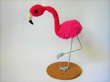 Rosa gehäkelter Amigurumi‑Flamingo mit gebogenem Hals auf runder Korkunterlage, ein Bein hochgezogen