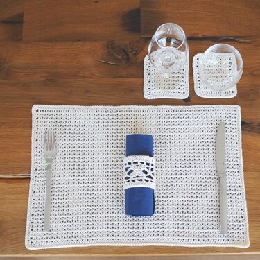 Beiges gehäkeltes Platzset mit zwei Untersetzern, blau gerollte Serviette, Besteck und Gläser auf Holztisch