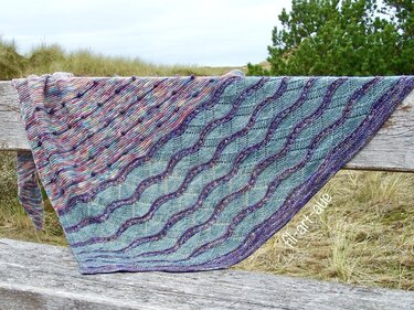 Handgestricktes Dreieckstuch in Türkis und mehrfarbigem Verlaufsgarn mit Wellenmuster, auf einer Holzbank vor Dünengras drapiert