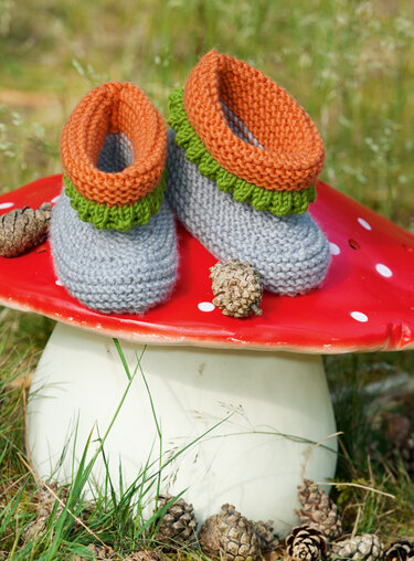Zwei handgestrickte Babyschuhe in Grau, Orange und Grün auf einem roten Deko-Pilz mit Kiefernzapfen.