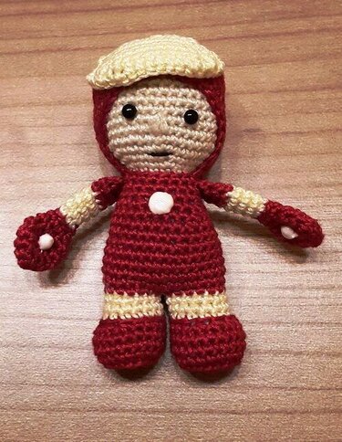 Gehäkelte Amigurumi-Puppe in Rot und Beige mit beiger Mütze, schwarzen Augen und weißen Akzenten auf Holzoberfläche.
