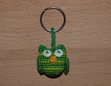 Gehäkelter grün-gelb gestreifter Eulen-Schlüsselanhänger mit weißen Augen und orangem Schnabel an Metallring auf Holzuntergrund