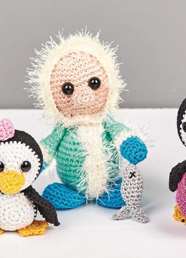 Gehäkelte Figur in türkisem Winteroutfit mit flauschiger Kapuze hält einen kleinen grauen Fisch; links und rechts zwei gehäkelte Pinguine auf weißem Untergrund.