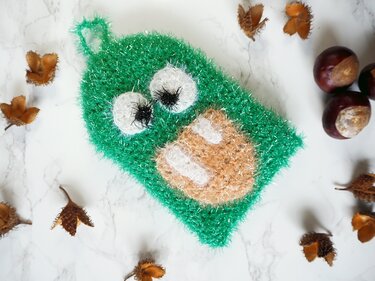 Grünes, flauschiges gehäkeltes Spültuch in Monsterform mit zwei weißen Augen und braunem Mundbereich, liegend auf hellem Marmoruntergrund mit Kastanien und getrockneten Blättern.