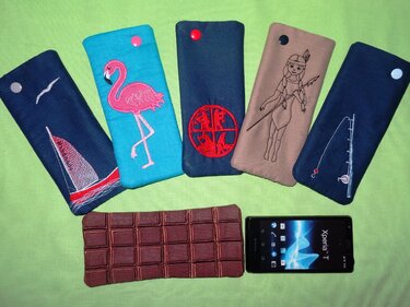 Sechs bestickte ITH-Handytaschen auf grünem Hintergrund mit Smartphone als Größenvergleich