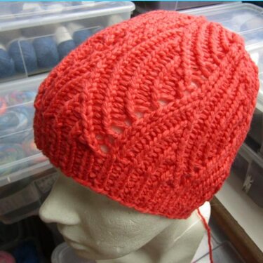 Korallrote Beanie mit Rippbündchen und Ajourstreifen auf Schaufensterkopf, Arbeitsfaden sichtbar