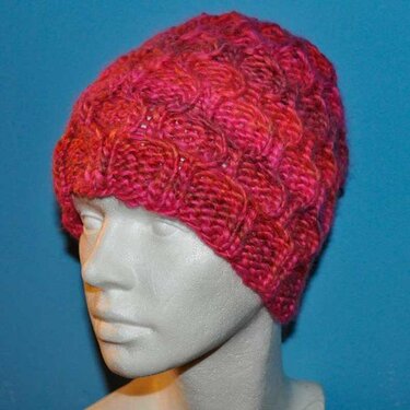 Gestrickte pinke Damenmütze mit Zopfmuster auf Schaufensterkopf vor blauem Hintergrund