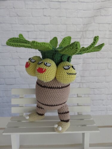 Gehäkelte dreiköpfige Pflanzenfigur (Amigurumi) mit gelben Köpfen und grünen Blättern auf weißer Holzbank vor heller Ziegelwand