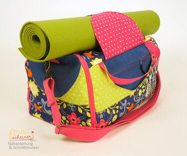 Bunte genähte Stofftasche mit Blumen- und Punktmustern, pinken Trägern und einer grünen eingerollten Yogamatte obenauf.