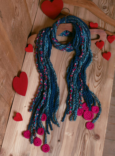 Gehäkelter blau-pinker Schal mit langen Fransen und pinken Rosetten auf Holzbrett mit roten Dekoherzen