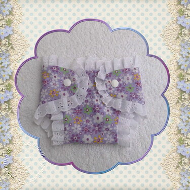 Lila Puppenwindel mit Blumenmuster, weißen Spitzenrüschen und zwei weißen Druckknöpfen auf hellem Stoff.