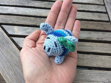 Kleine blau-grüne Amigurumi‑Schildkröte in einer Hand vor Holzlatten