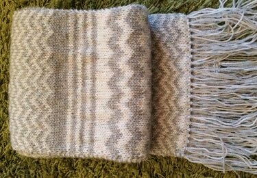 Gestrickter Schal in Grau und Weiß mit Zickzackmuster und Fransen auf grünem Untergrund