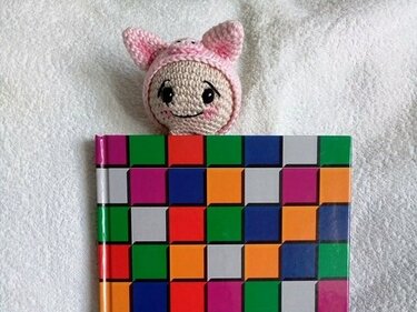Gehäkeltes Lesezeichen: rosa Katzenkopf über buntem Buch auf weißem Stoff